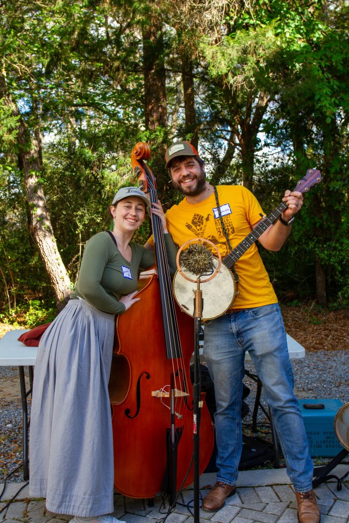 two people stand with instruments behind a gold microphone. one holds a banjo and the other stands with an upright bass